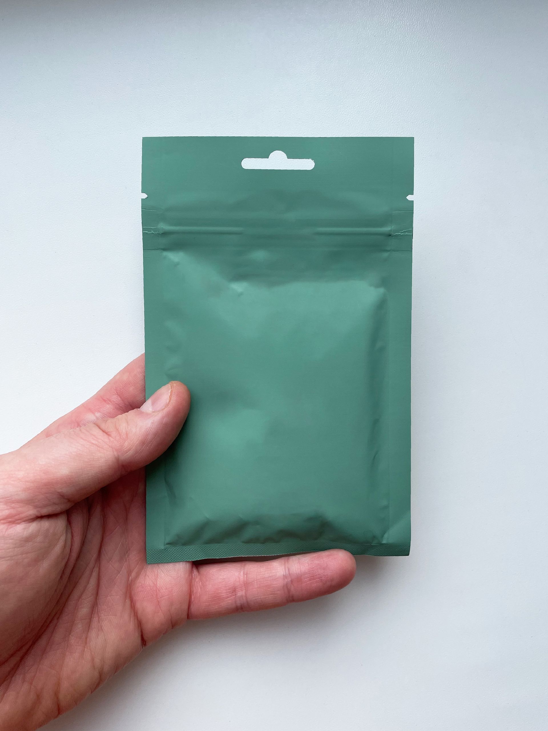 A green resealable pouch held by a hand on a neutral backdrop.